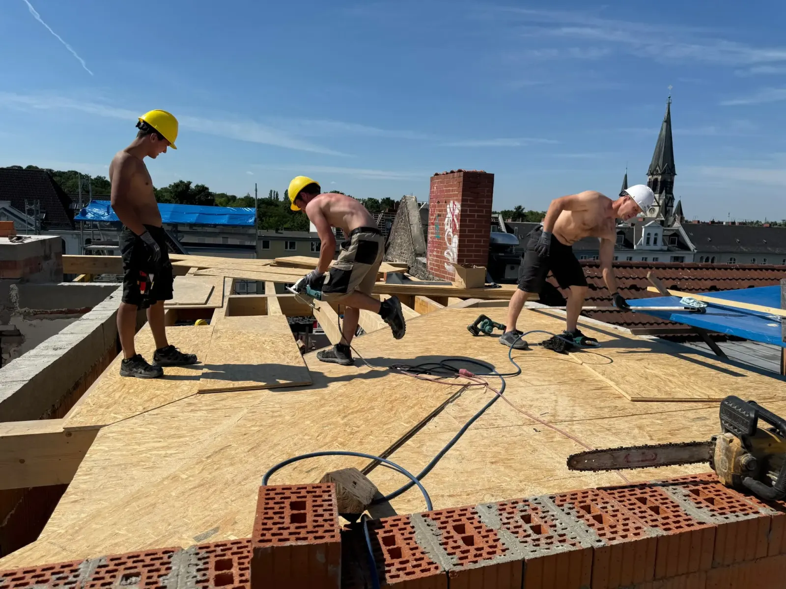 Drei Arbeiter verlegen OSB-Platten auf einem neuen Dachstuhl, Berliner Kirchturm im Hintergrund